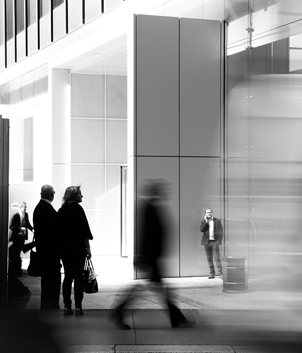 Scène urbaine en noir et blanc devant un bâtiment moderne vitré, avec des passants en mouvement flou et une personne immobile près de l’entrée.