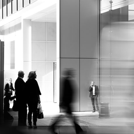 Scène urbaine en noir et blanc devant un bâtiment moderne vitré, avec des passants en mouvement flou et une personne immobile près de l’entrée.